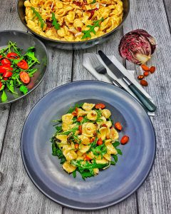 Auf einem grauen Teller angerichtet Italienische Nudeln - Orecchiette mit Macarpone und Rucola. Im Hintergrund eine Pfanne mit demselben Gericht und neben dem Teller eine Schale mit grünem Salat.