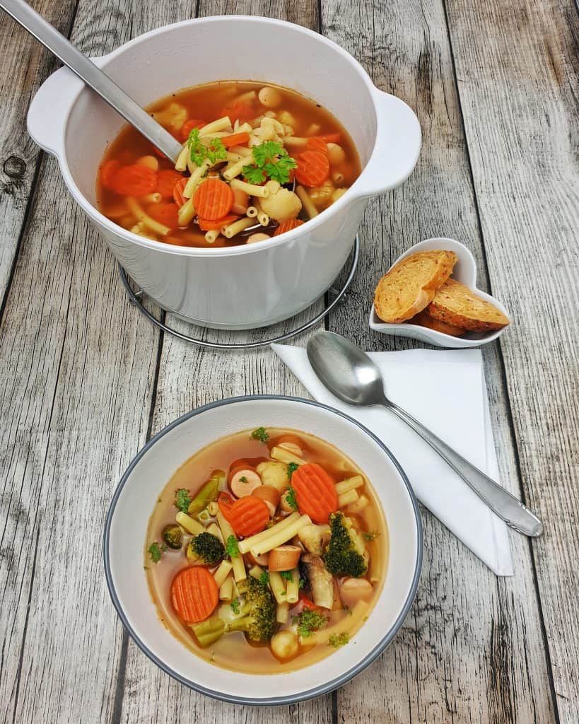 In einer weißen Schale angerichtet eine Gemüse-Nudel-Suppe mit Bockwurst. Im Hintergrund ein weißer Keramik-Topf mit Suppe.
