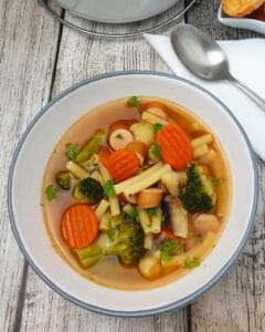 In einer weißen Schale angerichtet eine Gemüse-Nudel-Suppe mit Bockwurst. Im Hintergrund ein weißer Keramik-Topf mit Suppe.