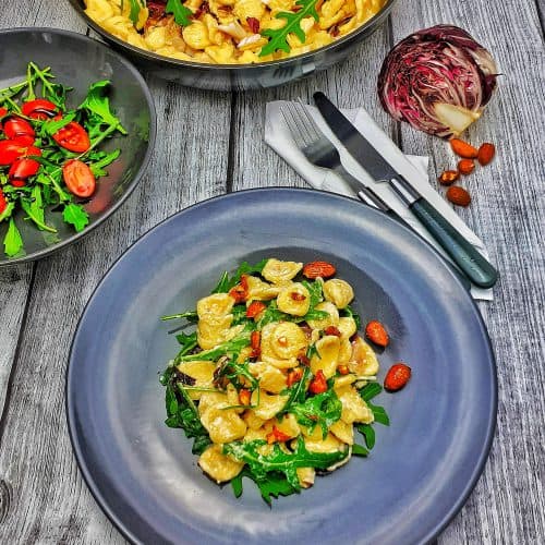 Auf einem grauen Teller angerichtet Italienische Nudeln - Orecchiette mit Macarpone und Rucola. Im Hintergrund eine Pfanne mit demselben Gericht und neben dem Teller eine Schale mit grünem Salat.
