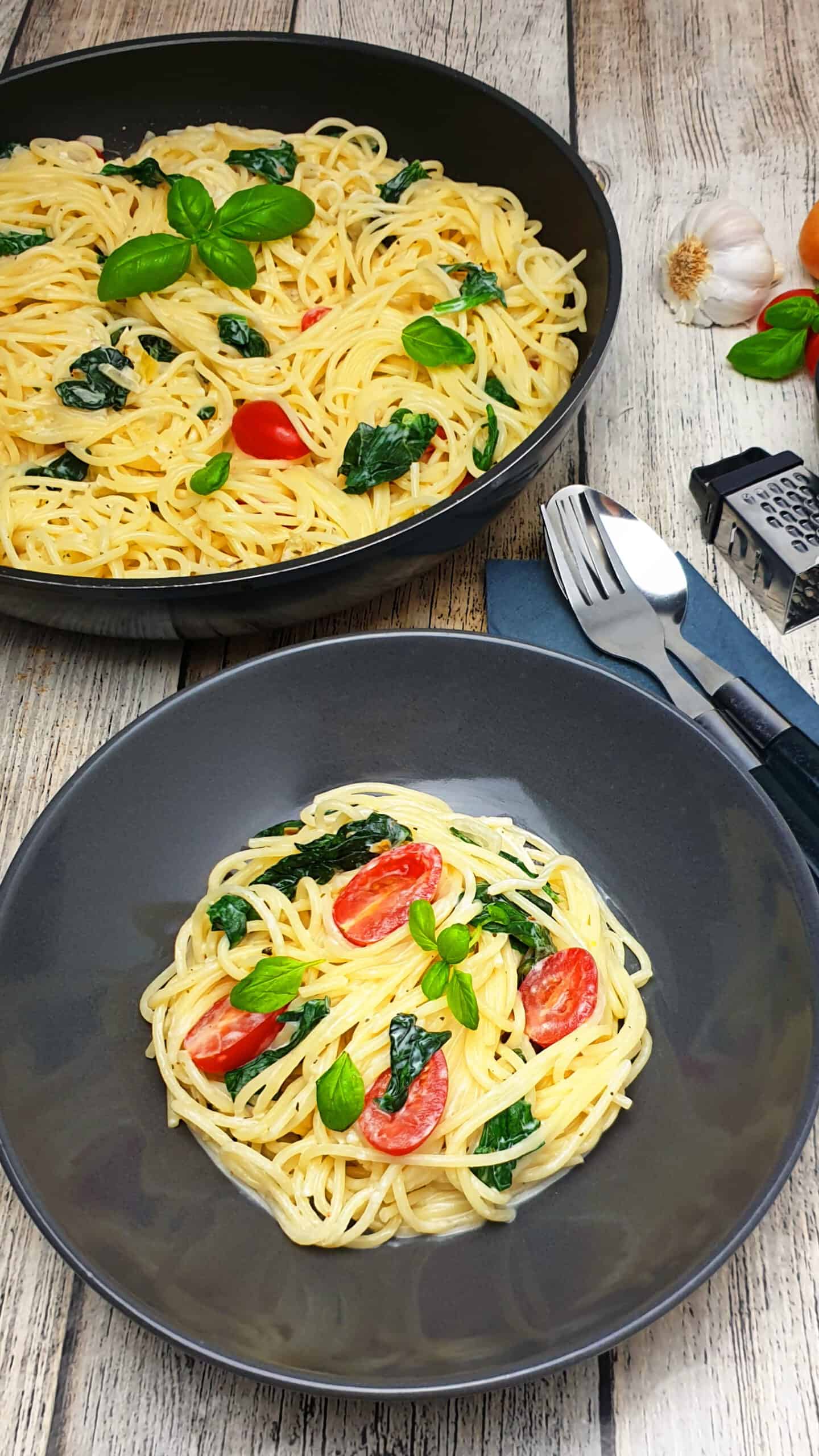 In einer grauen Schale ein Spaghetti Gericht mit Spinat und Tomate. Im Hintergrund Deko. 