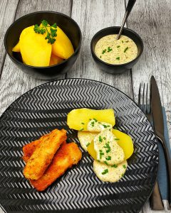 Auf einem schwarzen Teller angerichtet 3 selbstgemachte Fischstäbchen mit Salzkartoffeln und einer Dill-Senf-Soße. Im Hintergrund eine Schale mit Kartoffeln und eine Schale mit Dill-Senf-Soße.