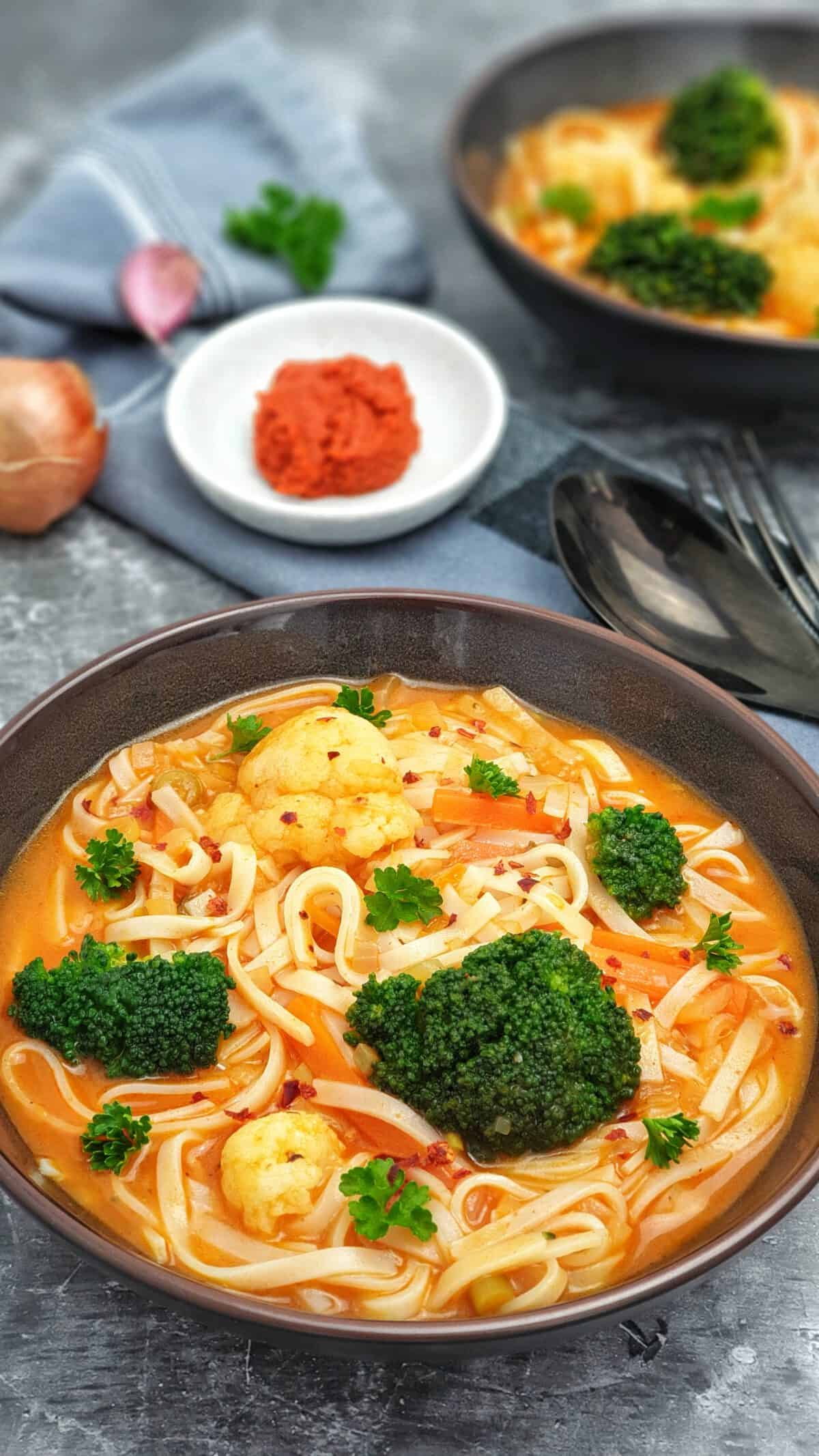 In einer braunen Schale eine rote Currysuppe mit Glasnudeln, Brokkoli und Blumenkohl. Im Hintergrund Deko. 