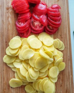 Auf einem Schneidebrett liegen in Scheiben geschnittene Tomaten und Kartoffeln für den Kartoffel Frikadellen Auflauf.