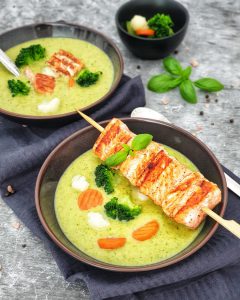 Brokkoli Creme Suppe mit einem Lachsspieß in einer braunen Schale serviert. Der Lachsspieß ist angebraten und auf dem Schälchen drapiert.
