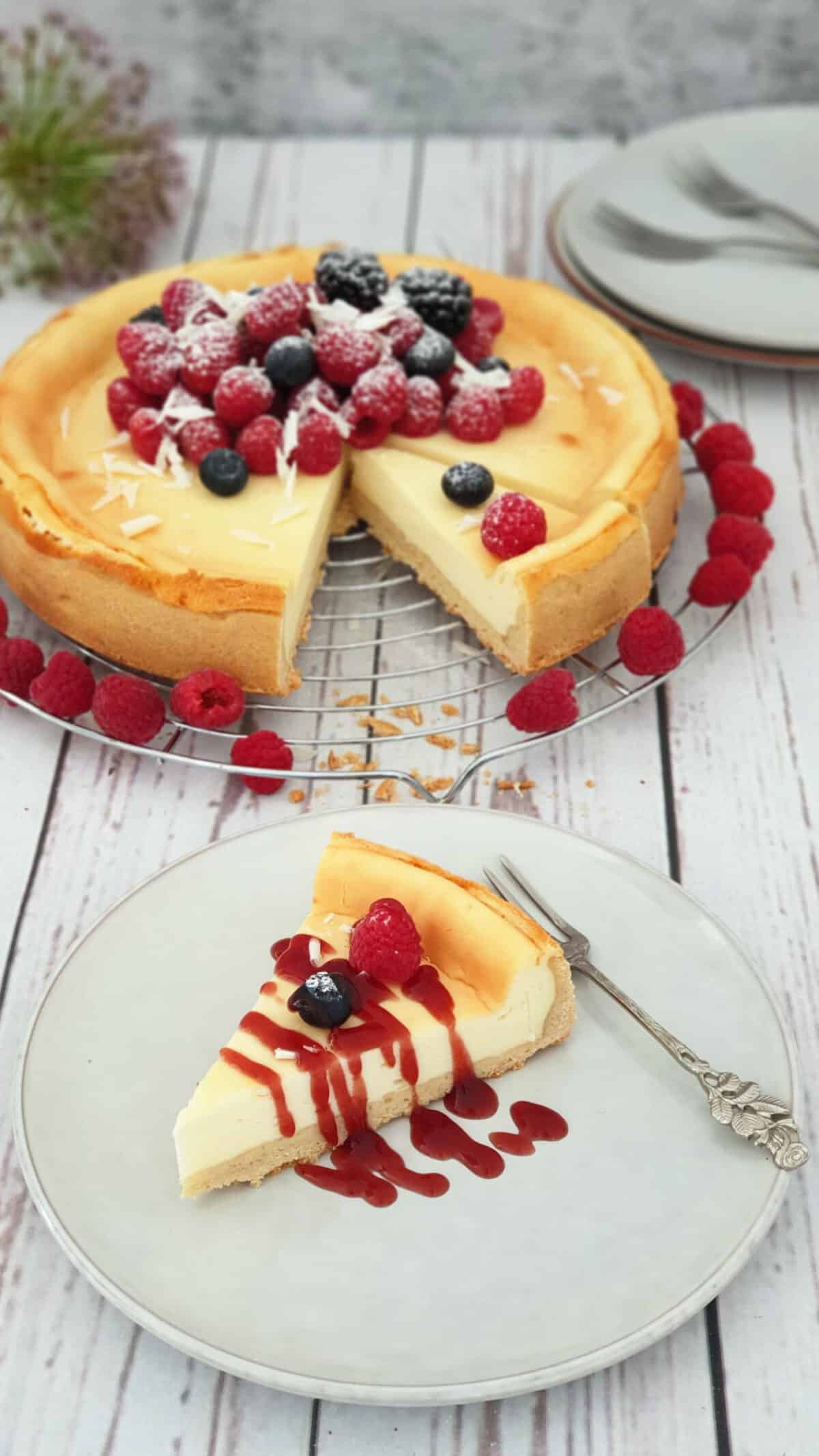 Auf einem hellen Teller ein Stück Cheesecake mit Fürchten und Beeren. Im Hintergrund ein Käsekuchen auf einem Kuchenrost. 