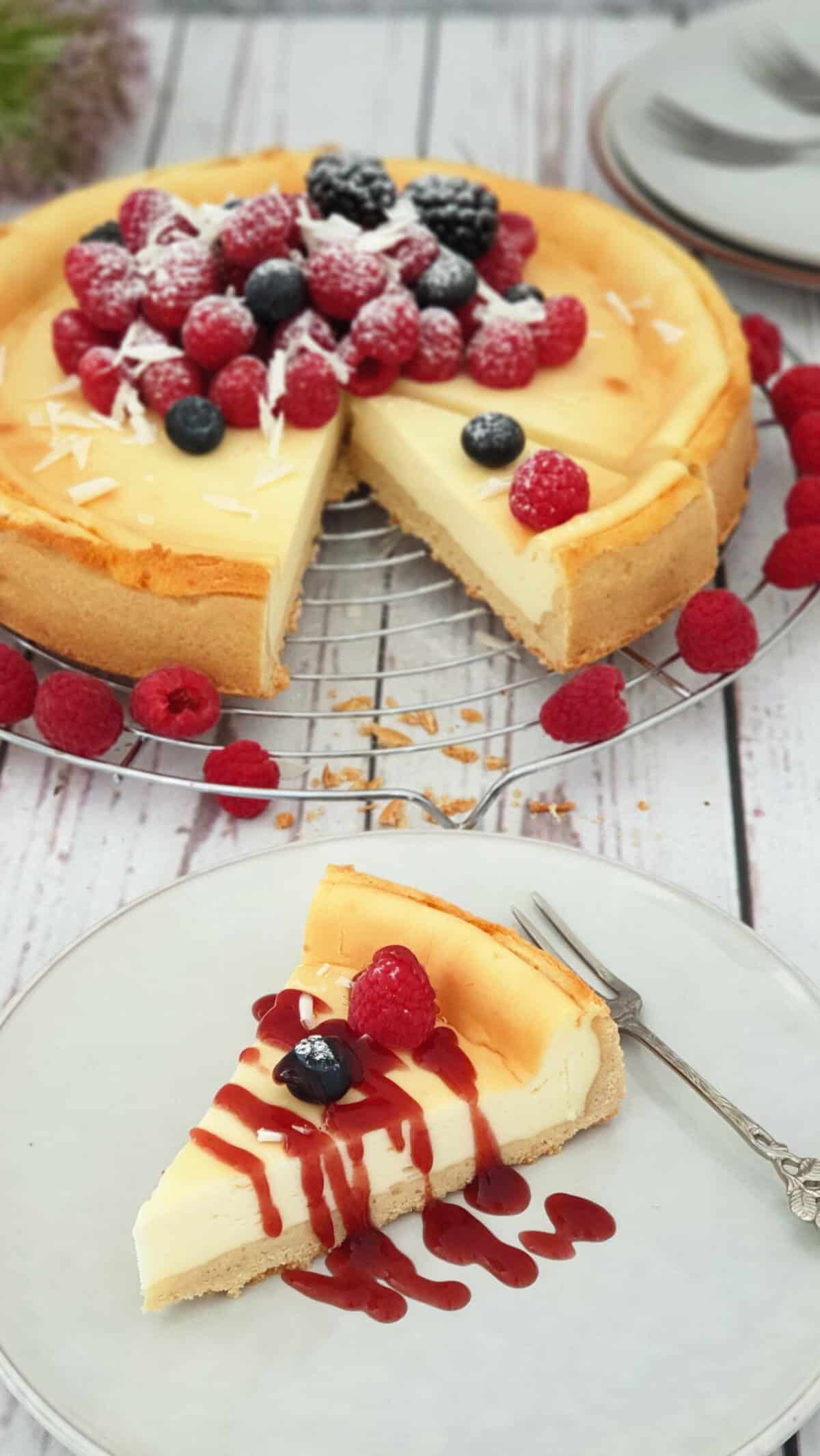 Auf einem hellen Teller ein Stück Cheesecake mit Fürchten und Beeren. Im Hintergrund ein Käsekuchen auf einem Kuchenrost.