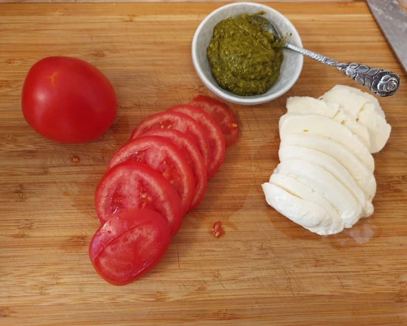 Auf einem Schneidebrett Tomaten und Mozzarella Scheiben, daneben eine Schale mit grünem Pesto.