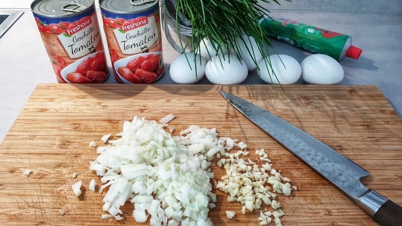 Auf einem Schneidebrett klein geschnittene Zwiebeln. Im Hintergrund Kräuter, Tomaten und Eier.