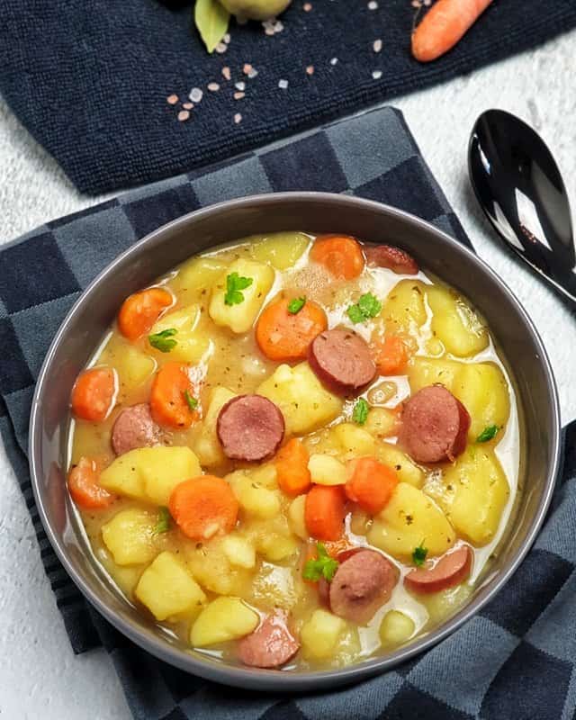 Ein Kartoffel Eintopf mit Möhren und Rindswurst in einer grauen Schale angerichtet und auf einem Küchenhandtuch fotografiert