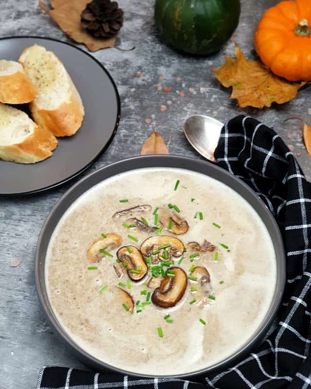 Pilz-Creme-Suppe dekorativ angerichtet in einer grauen Schale, mit ein paar Pilzen in der Mitte und frischem Schnittlauch angerichtet. Im Hintergrund herbstliche Dekoration und ein Kräuter Baguette