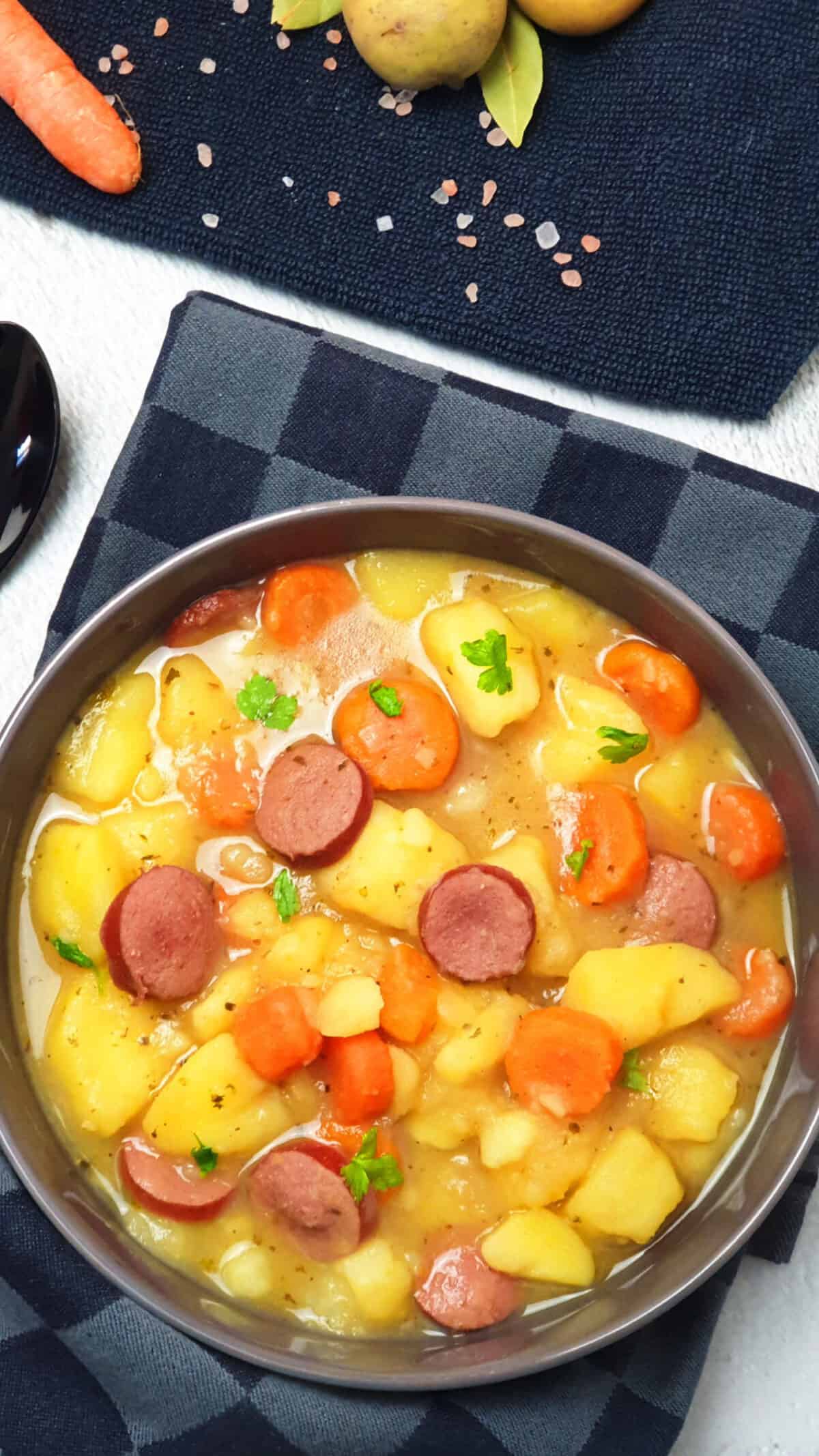 In einer grauen Schale ein Kartoffel-Möhren-Eintopf mit einer Frankfurter Rindswurst. Im Hintergrund Deko.