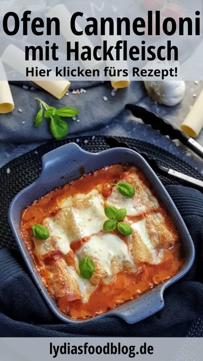 In einer grauen Auflaufform zu sehen, gefüllte Cannelloni mit Hackfleisch und Mozzarella aus dem Ofen in einer Tomatensoße. Im Hintergrund dekorativ Cannelloni Nudeln, Tomaten, Knoblauch und Basilikum drapiert. Neben der Auflaufform eine Küchenzange.