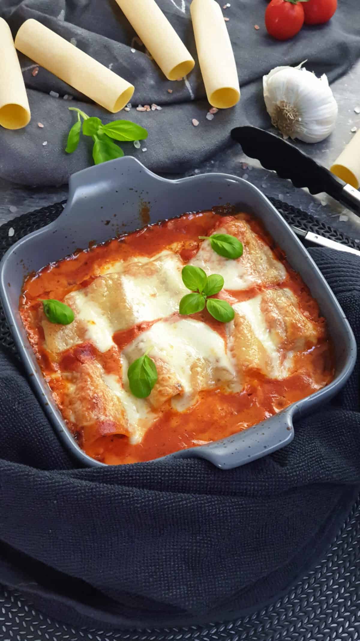 In einer grauen Auflaufform gefüllte Cannelloni mit Hackfleisch in Tomatensoße. Im Hintergrund Deko.