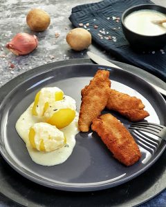 3 selbstgemachte Fischstäbchen mit 3 kleinen Kartoffeln auf einem grauen Teller. Unter und auf den Kartoffeln eine Dill-Senf-Soße. Im Hintergrund Deko