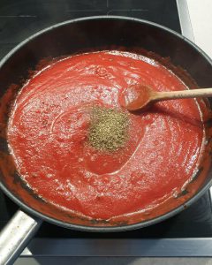 In einer schwarzen Pfanne kocht eine Tomatensoße, ein Rührlöffel ist im oberen Rand des Bildes zu erkennen. In der Pfannenmitte sieht man getrocknete Kräuter, die gerade erst in die Pfanne gegeben wurden.