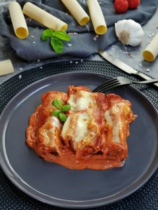 Auf einem dunklen Teller zu sehen, 3 gefüllte Cannelloni in einer Tomatensoße und mit Mozzarella überbacken. Im Hintergrund auf einem Handtuch dekorativ Cannelloni Nudeln, Tomaten, Knoblauch und Basilikum drapiert. Neben dem Teller zu sehen eine dunkle Gabel und ein dunkles Messer in mattschwarz. Fertig sind die Cannelloni mit Hackfleisch und Mozzarella aus dem Ofen.