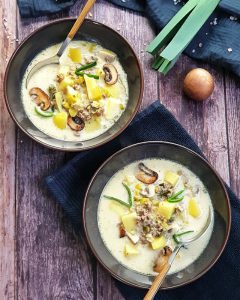 Zwei braune Suppenschälchen, gefüllt mit einer Käse-Lauch-Suppe. Im Hintergrund sieht man eine Stange Lauch, eine Zwiebel und Champignons, dekorativ auf einem Küchenhandtuch fotografiert.