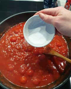 Zu sehen ist wie eine Prise Zucker zur Tomatensoße dazu gegeben wird.