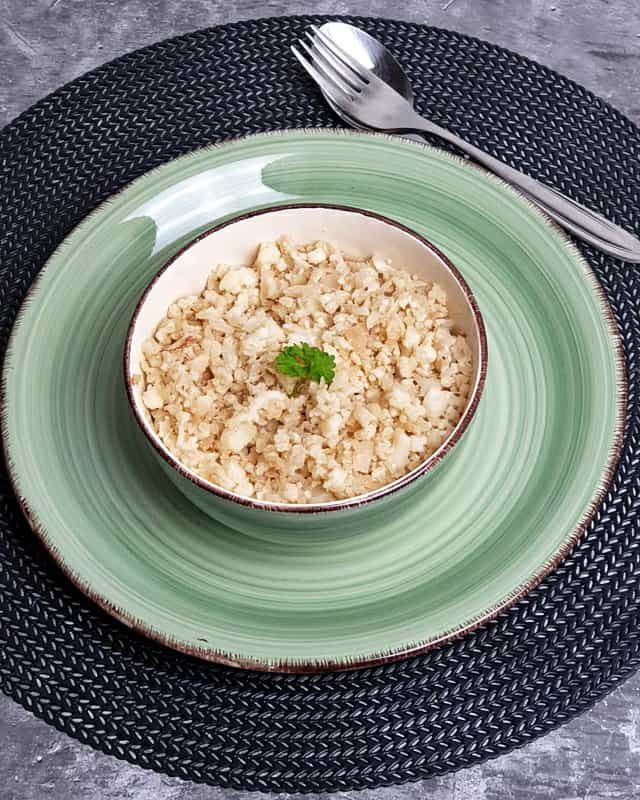 In einer hellen Schale auf einem grünen Teller angebratenen Blumenkohlreis, schneller Low Carb Reis mit etwas Petersilie als Deko.