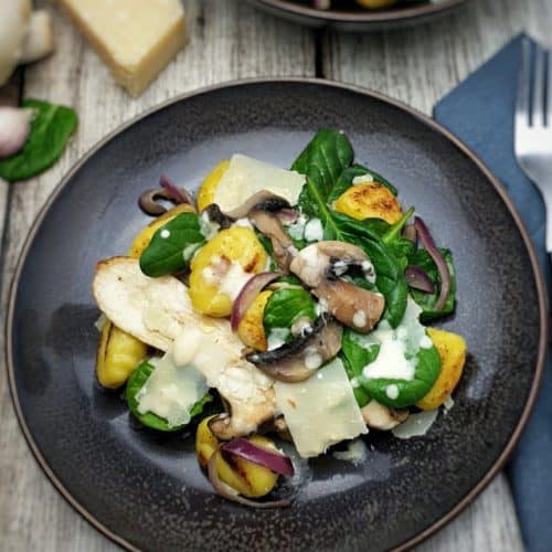 Auf einem braunen Teller angerichtet, ein Gnocchi Salat mit angebratenen Pilzen, frischem Spinat und gehobeltem Parmesan.