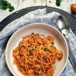 In einer weißen, runden Schale angerichtet Spaghetti mit Shrimps und Tomatensoße. Die Spaghetti sind mit Petersilie und Parmesan bestreut. Neben dem Teller liegt ein schwarzes Besteck. Im Hintergrund Deko.