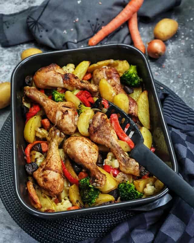In einer schwarzen Auflaufform sieht man das fertige Hähnchenschenkel mit Gemüse und Kartoffeln aus dem Ofen. Im Hintergrund sind 2 Möhren, ein paar Kartoffeln und eine Zwiebel zu erkennen.