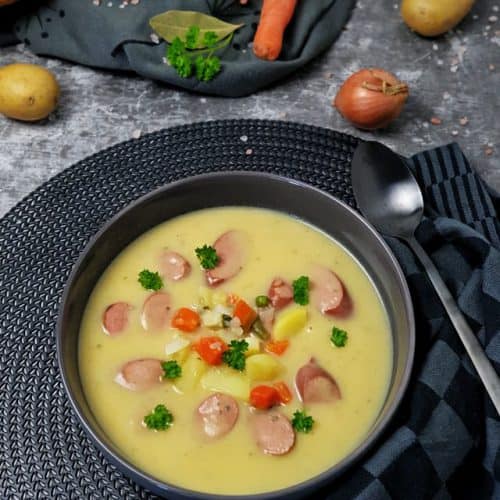 In einer grauen Schale dekorativ angerichtet eine cremige Kartoffelsuppe mit Würstchen sowie Kartoffel und Gemüsestücken. Im Hintergrund auf einem Handtuch liegt ein Baguette, eine Möhre und Kartoffeln.