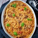 Spaghetti Auflauf mit Hackfleisch in einer weißen Auflaufform mit Basilikum Blätter bestreut. Um die Auflaufform ein dunkles Küchenhandtuch. Daneben auf einem weiteren Küchentuch eine Spaghetti Zange.