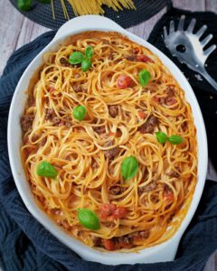 Spaghetti Auflauf mit Hackfleisch in einer weißen Auflaufform mit Basilikum Blätter bestreut. Um die Auflaufform ein dunkles Küchenhandtuch. Daneben auf einem weiteren Küchentuch eine Spaghetti Zange.