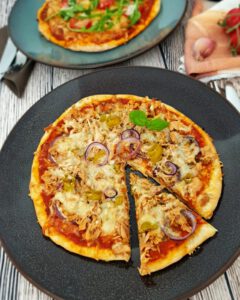 Auf einem dunklen großen Teller eine Dinkel Pizza mit Thunfisch, Zwiebeln und Käse.