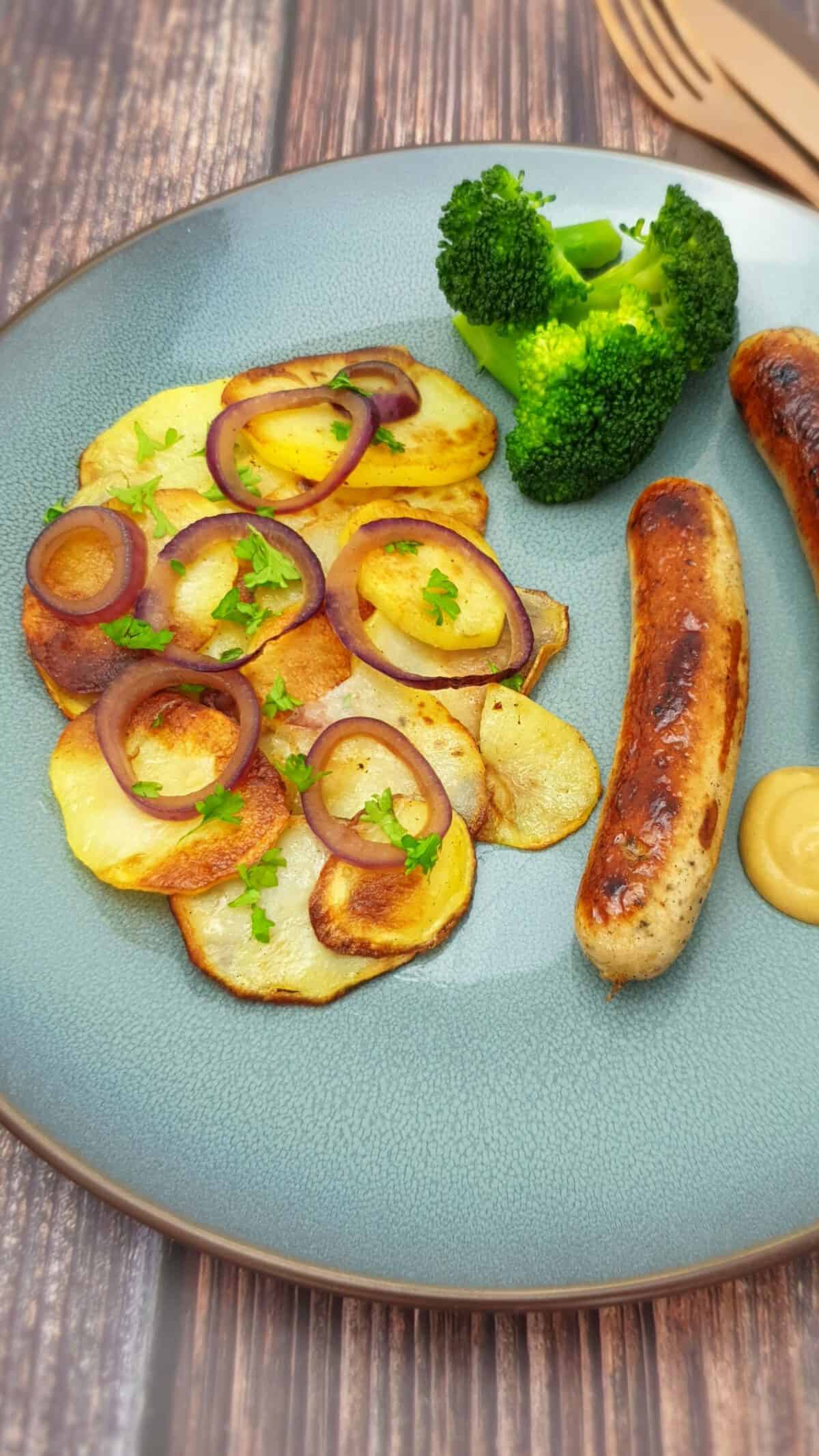Knusprige Bratkartoffeln mit roten Zwiebel und einer Bratwurst auf einem blauen Teller.
