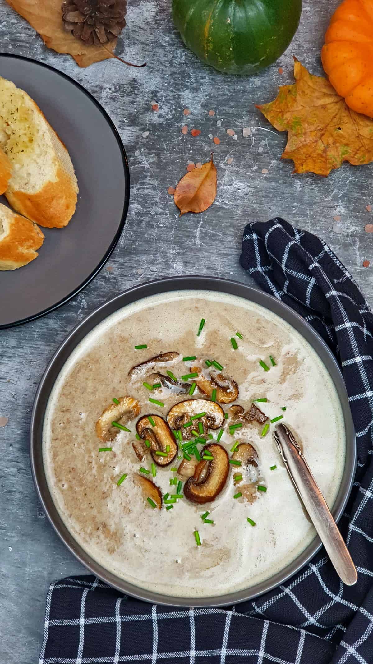 Pilz Creme Suppe mit Champignons in einer grauen Schale serviert. Mit Schnittlauch bestreut. Im Hintergrund Herbst Deko.