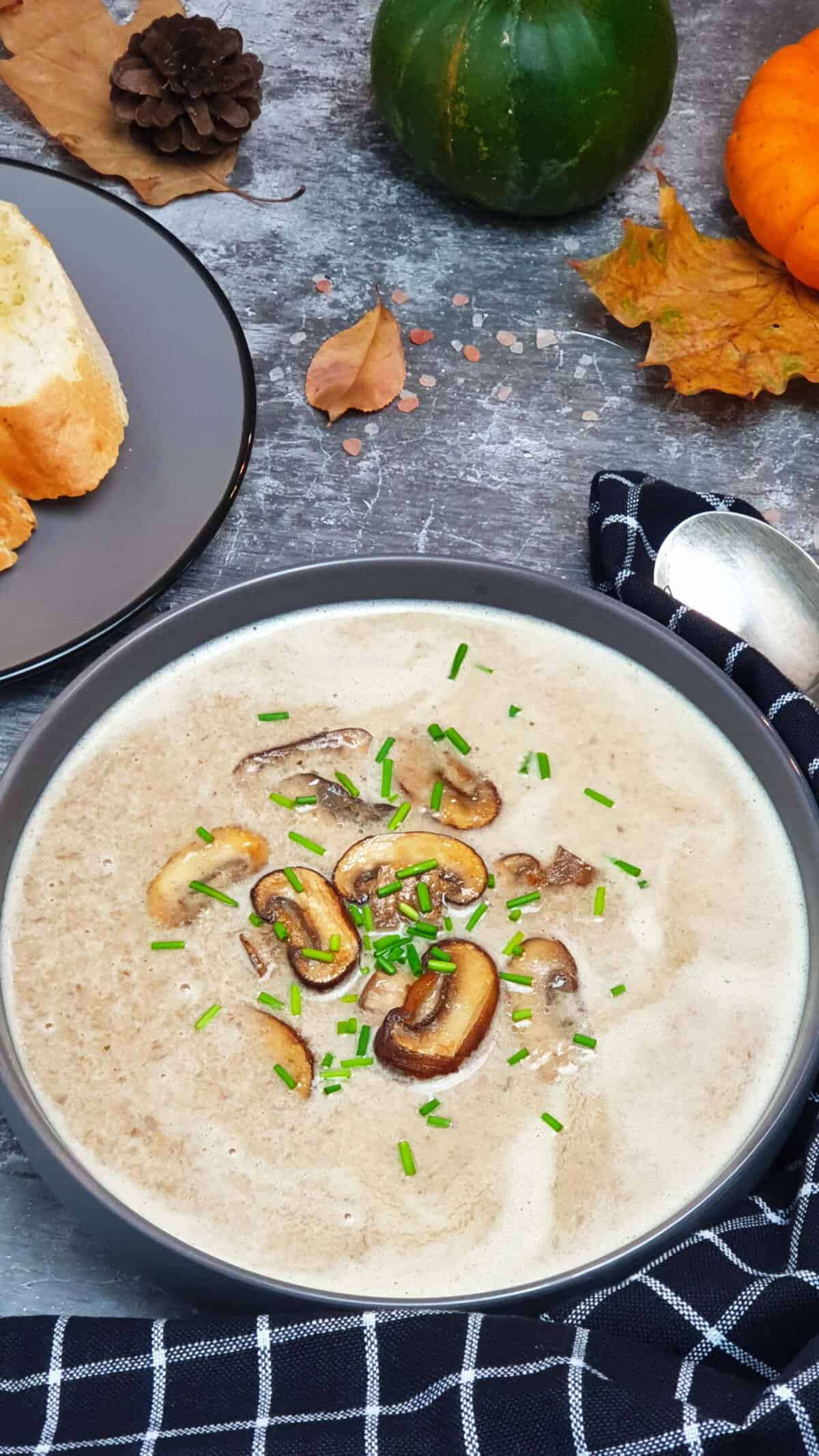 Pilz Creme Suppe mit Champignons in einer grauen Schale serviert. Mit Schnittlauch bestreut. Im Hintergrund Herbst Deko.