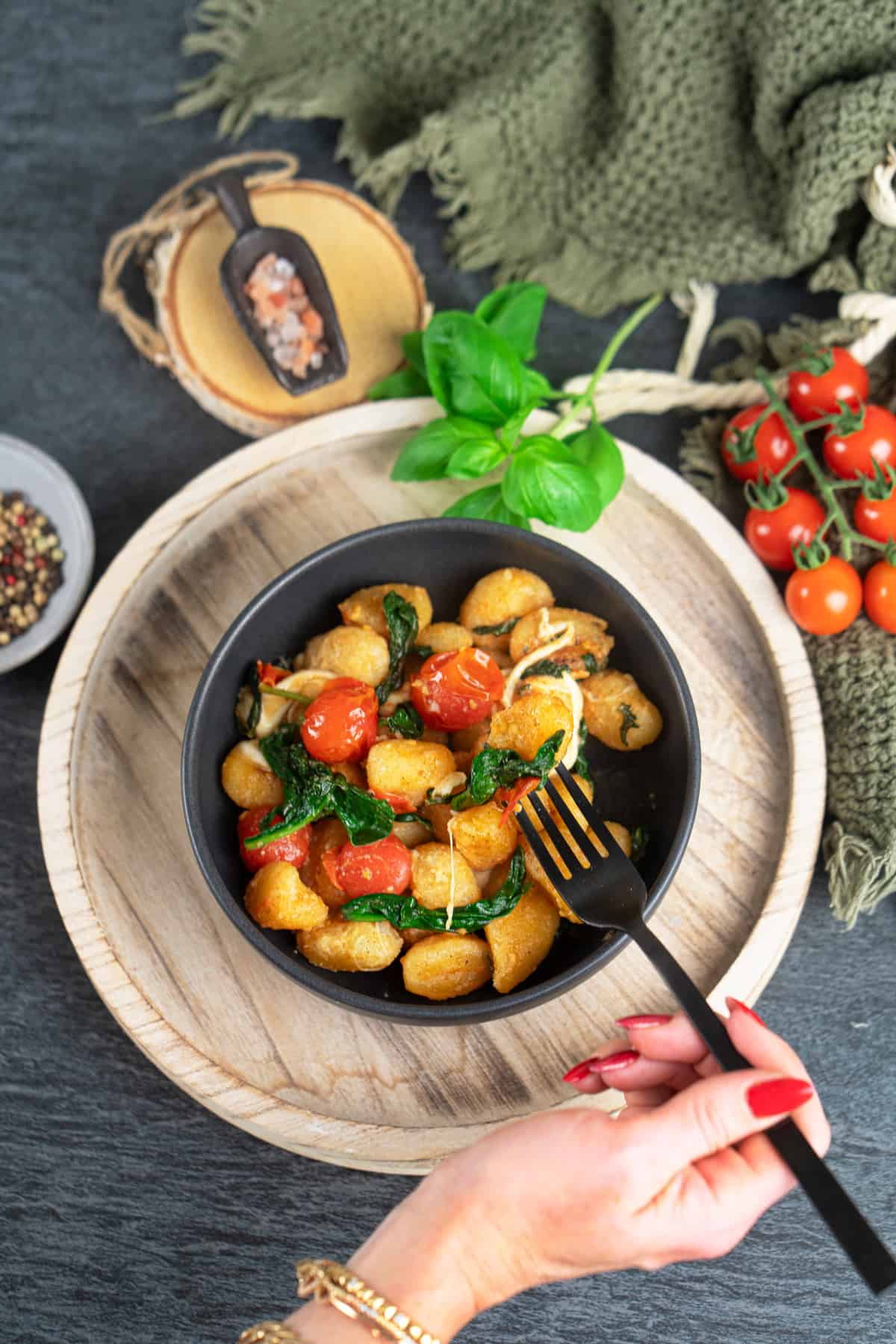 In einer schwarzen Schale angerichtet Gnocchi mit Tomaten, Spinat und Burrata. Eine Hand hält eine Gabel mit Gnocchi und Tomate. 